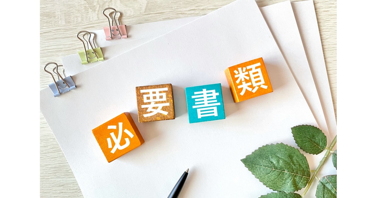 alt="重ねた白いA4用紙の中央に、オレンジ・茶・ターコイズの木製ブロックで『必 要 書 類』の4文字が斜めに並ぶ。左上にはパステルカラーのスマイルクリップ、右下にはアイビーの葉と黒ペンが添えられ、提出書類の準備をイメージした写真"