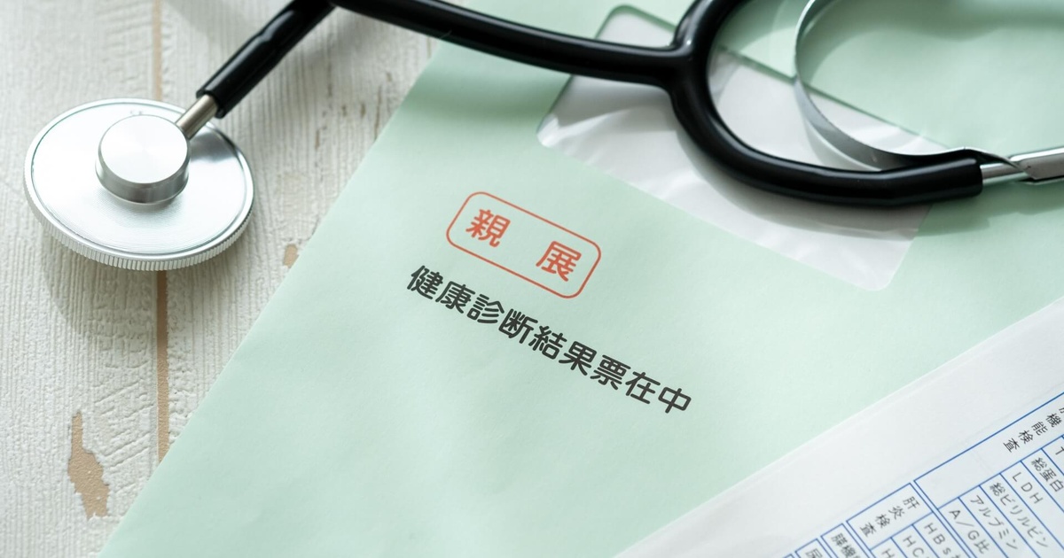 alt="木目テーブルの上に置かれた淡いグリーンの封筒には『親展 健康診断結果票在中』と印刷され、左上には銀色の聴診器、右下には検査項目が並ぶ用紙が覗き、健康診断の結果通知を連想させる写真"