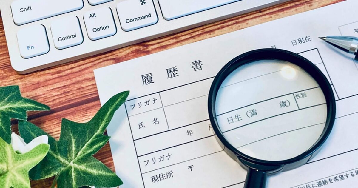alt="木目のデスクに置かれた履歴書を黒い虫眼鏡で確認する場面。左上に白いPCキーボード、左下にアイビーの葉、右上に銀色のボールペンが映り込み、就職・転職準備をイメージさせる写真"