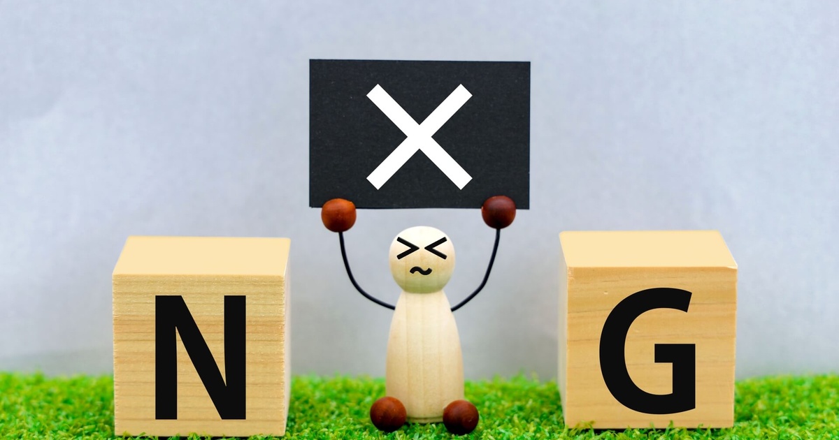 芝生の上に置かれた木製ブロック人形が苦い表情で黒いバツ印のパネルを掲げ、両脇にNとGの文字ブロックが並び『NG』を示す写真
