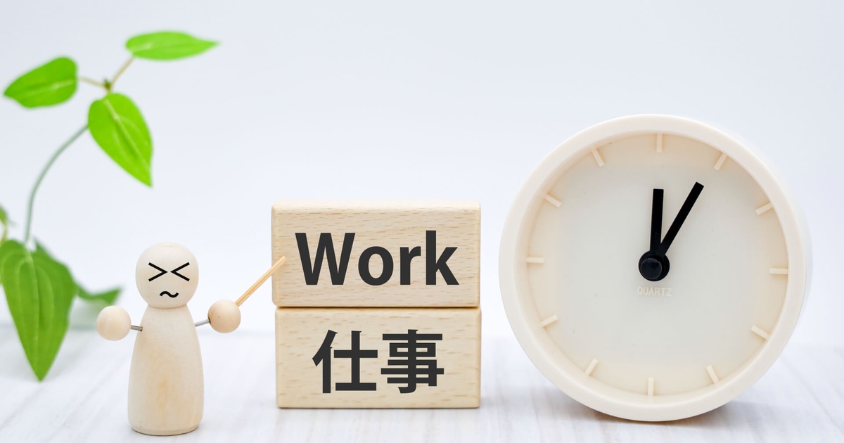 白い背景に木製ブロックで『Work』『仕事』と書かれ、隣に怒った表情の木製人形が指し棒を構え、右側には2時を指す白い時計が置かれた労働をイメージした写真