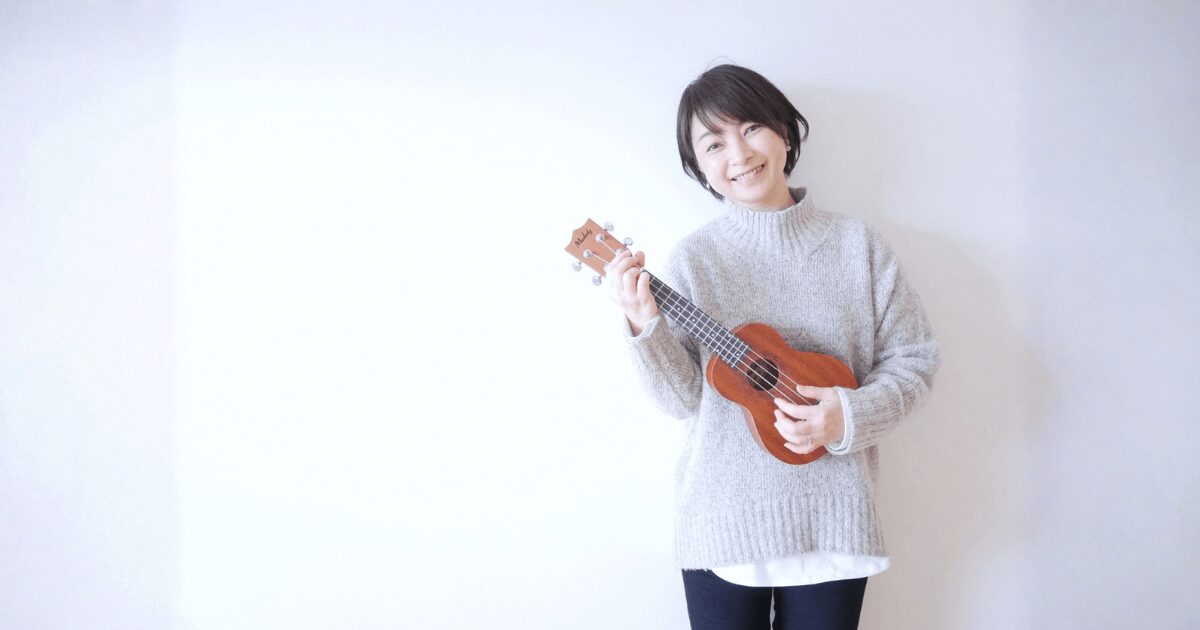 ウクレレを手にした女性が笑顔で立っている写真。 楽器演奏を楽しむ穏やかな雰囲気を伝えるイメージ。
